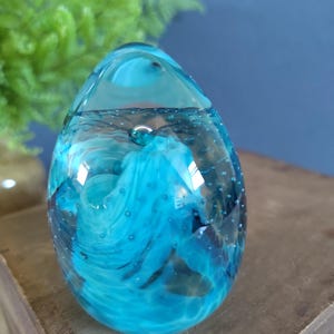 May include: A clear, egg-shaped glass paperweight with swirling blue and turquoise patterns. The glass contains small air bubbles, adding texture. The paperweight sits on a wooden surface, with a blurred green plant in the background.
