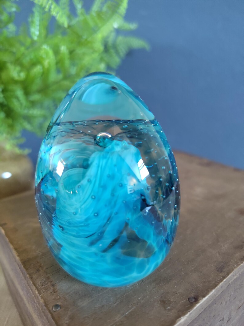 May include: A clear, egg-shaped glass paperweight with swirling blue and turquoise patterns. The glass contains small air bubbles, adding texture. The paperweight sits on a wooden surface, with a blurred green plant in the background.