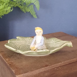 Puede incluir: Figura de cerámica de un gnomo sentado con sombrero amarillo y chaqueta azul, sosteniendo un objeto amarillo, colocado en una fuente verde con forma de hoja. La fuente está sobre una superficie de madera.