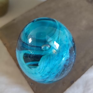 May include: A clear, round glass paperweight with a vibrant turquoise and white swirl design. The glass has small air bubbles throughout, adding texture. The paperweight sits on a dark, textured surface.