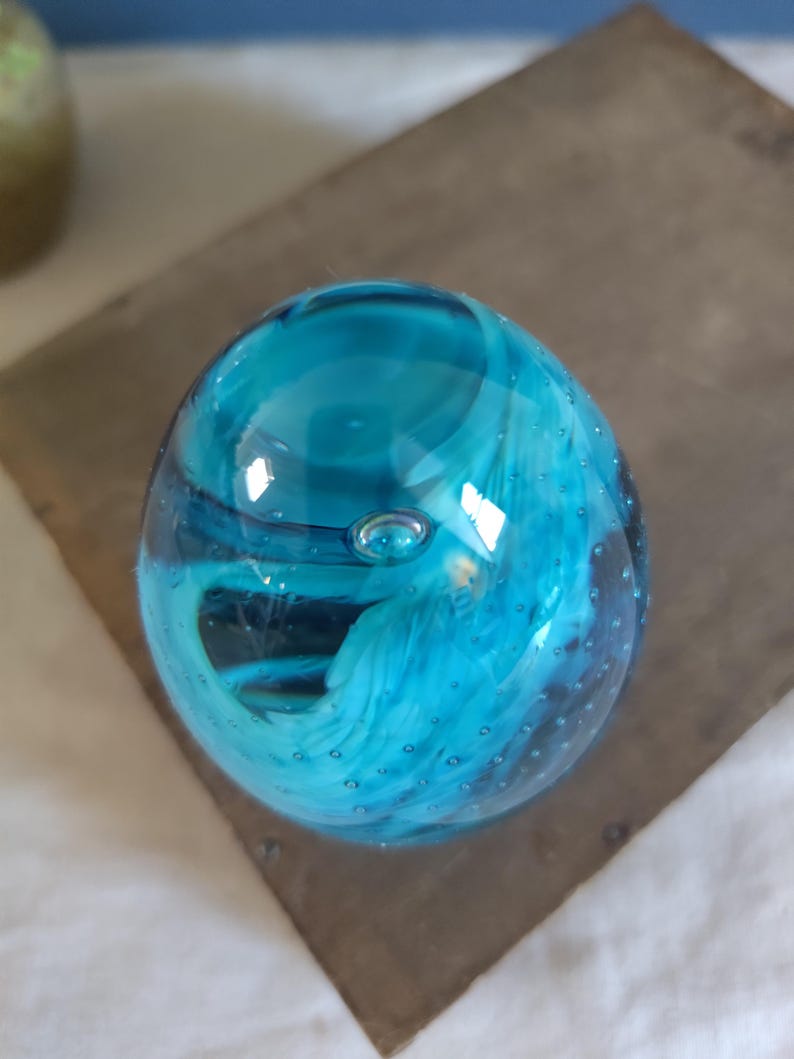 May include: A clear, round glass paperweight with a vibrant turquoise and white swirl design. The glass has small air bubbles throughout, adding texture. The paperweight sits on a dark, textured surface.