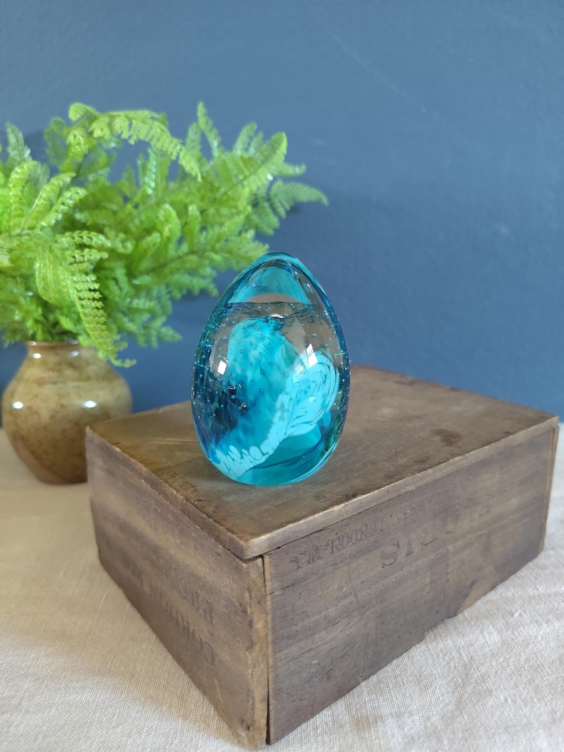 May include: A clear, egg-shaped glass paperweight with a vibrant turquoise interior design. The paperweight sits atop a weathered wooden box, with a fern in a ceramic vase in the background.