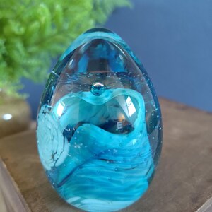 May include: A clear glass paperweight with a blue and white swirling design. The egg-shaped paperweight has a bubble at the top and is filled with shades of blue and white. The paperweight sits on a wooden surface.