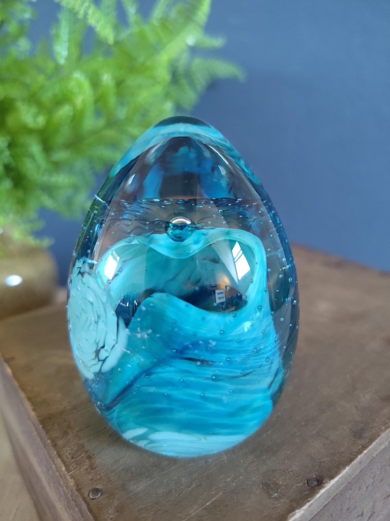 May include: A clear glass paperweight with a blue and white swirling design. The egg-shaped paperweight has a bubble at the top and is filled with shades of blue and white. The paperweight sits on a wooden surface.