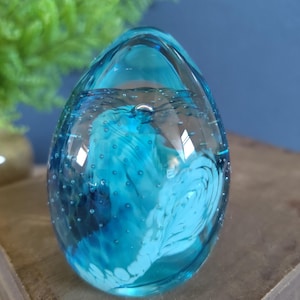 May include: A clear, egg-shaped glass paperweight with a vibrant blue and white swirl design inside. The glass is filled with tiny bubbles, adding texture. The paperweight sits on a wooden surface.