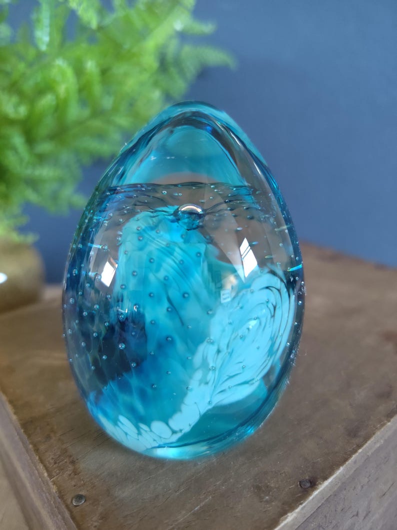 May include: A clear, egg-shaped glass paperweight with a vibrant blue and white swirl design inside. The glass is filled with tiny bubbles, adding texture. The paperweight sits on a wooden surface.