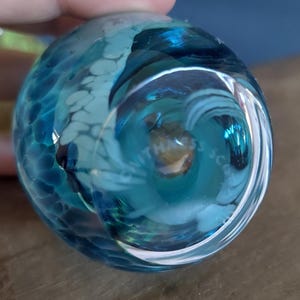 May include: A clear, round glass paperweight with swirling blue and white patterns. The glass has a textured appearance and a glossy finish. The bottom of the paperweight has the text "MASS SC".
