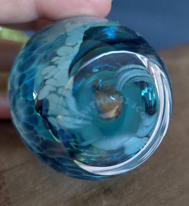 May include: A clear, round glass paperweight with swirling blue and white patterns. The glass has a textured appearance and a glossy finish. The bottom of the paperweight has the text "MASS SC".