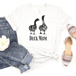 May include: A light grey t-shirt with the words "DUCK MOM" in black. The shirt features a graphic of two black geese with floral designs inside. The shirt is paired with blue jeans and patterned flats.