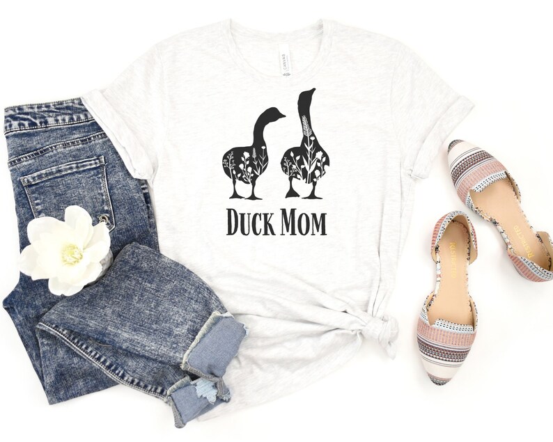May include: A light grey t-shirt with the words "DUCK MOM" in black. The shirt features a graphic of two black geese with floral designs inside. The shirt is paired with blue jeans and patterned flats.