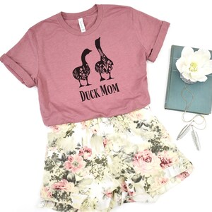 May include: A mauve t-shirt with the text "DUCK MOM" and a graphic of two geese filled with floral patterns. Paired with floral print shorts in cream, pink, and green. A book and jewelry are also in the image.