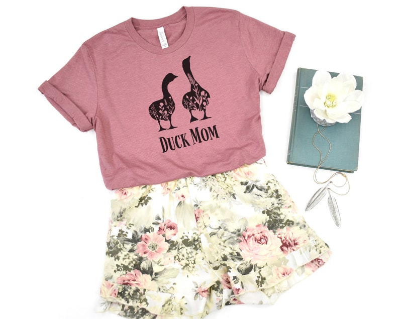 May include: A mauve t-shirt with the text "DUCK MOM" and a graphic of two geese filled with floral patterns. Paired with floral print shorts in cream, pink, and green. A book and jewelry are also in the image.