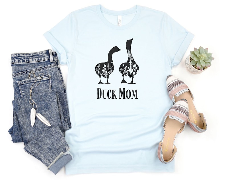 May include: Pale blue t-shirt featuring a black graphic of two geese with floral patterns. The words "Duck Mom" are printed beneath the geese. The image includes blue jeans and patterned shoes.