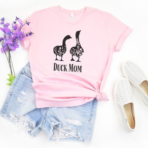 May include: A pale pink t-shirt with the text "DUCK MOM" in black. The shirt displays a graphic of two geese filled with floral patterns. The image includes denim shorts and white slip-on shoes.