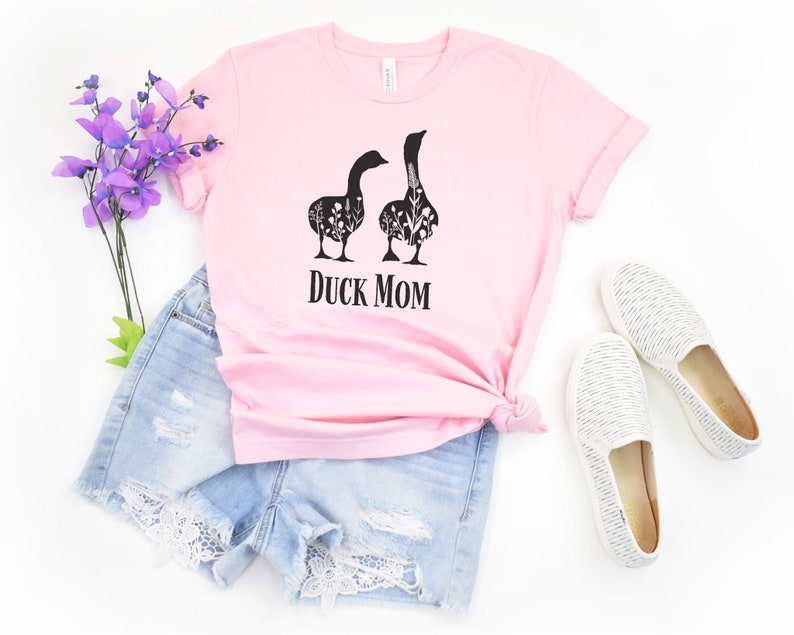 May include: A pale pink t-shirt with the text "DUCK MOM" in black. The shirt displays a graphic of two geese filled with floral patterns. The image includes denim shorts and white slip-on shoes.