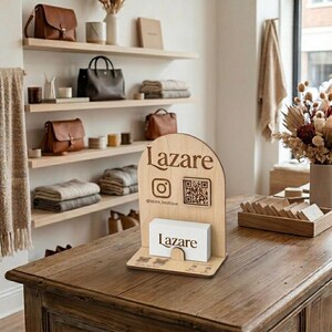 Puede incluir: Un expositor de madera con la palabra "Lazare" grabada, iconos de redes sociales y un código QR. Tarjetas de visita blancas con el logotipo "Lazare" están en el expositor. El expositor está sobre una mesa de madera.