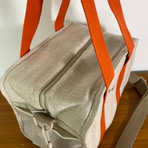 May include: A beige canvas tote bag with orange straps and a zipper closure. The bag has a mesh pocket on the side and a long strap for carrying it on the shoulder.