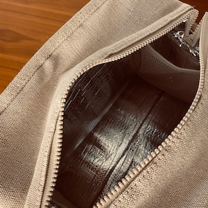May include: A beige canvas lunch bag with a zipper opening to reveal a silver insulated lining. The bag has a small mesh pocket inside.