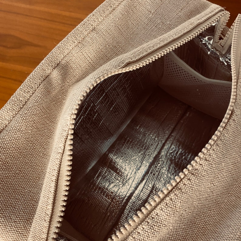 May include: A beige canvas lunch bag with a zipper opening to reveal a silver insulated lining. The bag has a small mesh pocket inside.
