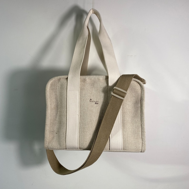 May include: A white canvas tote bag with a brown adjustable strap. The bag has two white straps and the text "The Well-Heeled Home" is printed on the front.