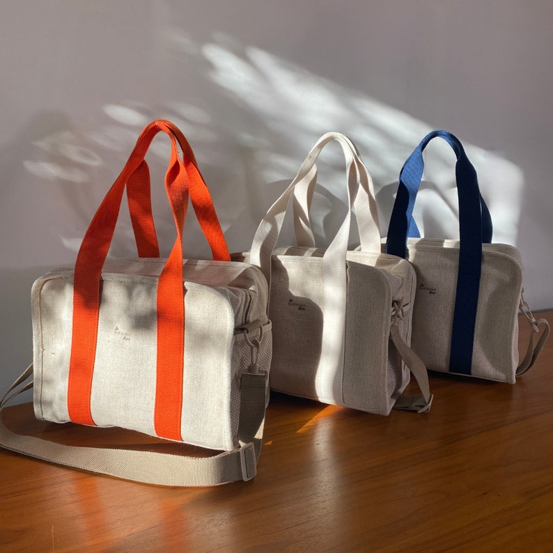 May include: Three canvas tote bags with different colored straps. The bag on the left has orange straps, the middle bag has beige straps, and the bag on the right has navy blue straps.