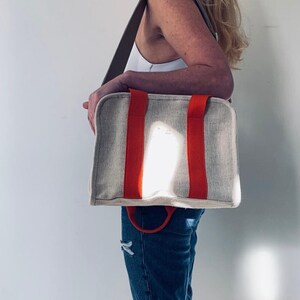 May include: A woman is carrying a beige canvas tote bag with orange straps. The bag has a top handle and a shoulder strap.