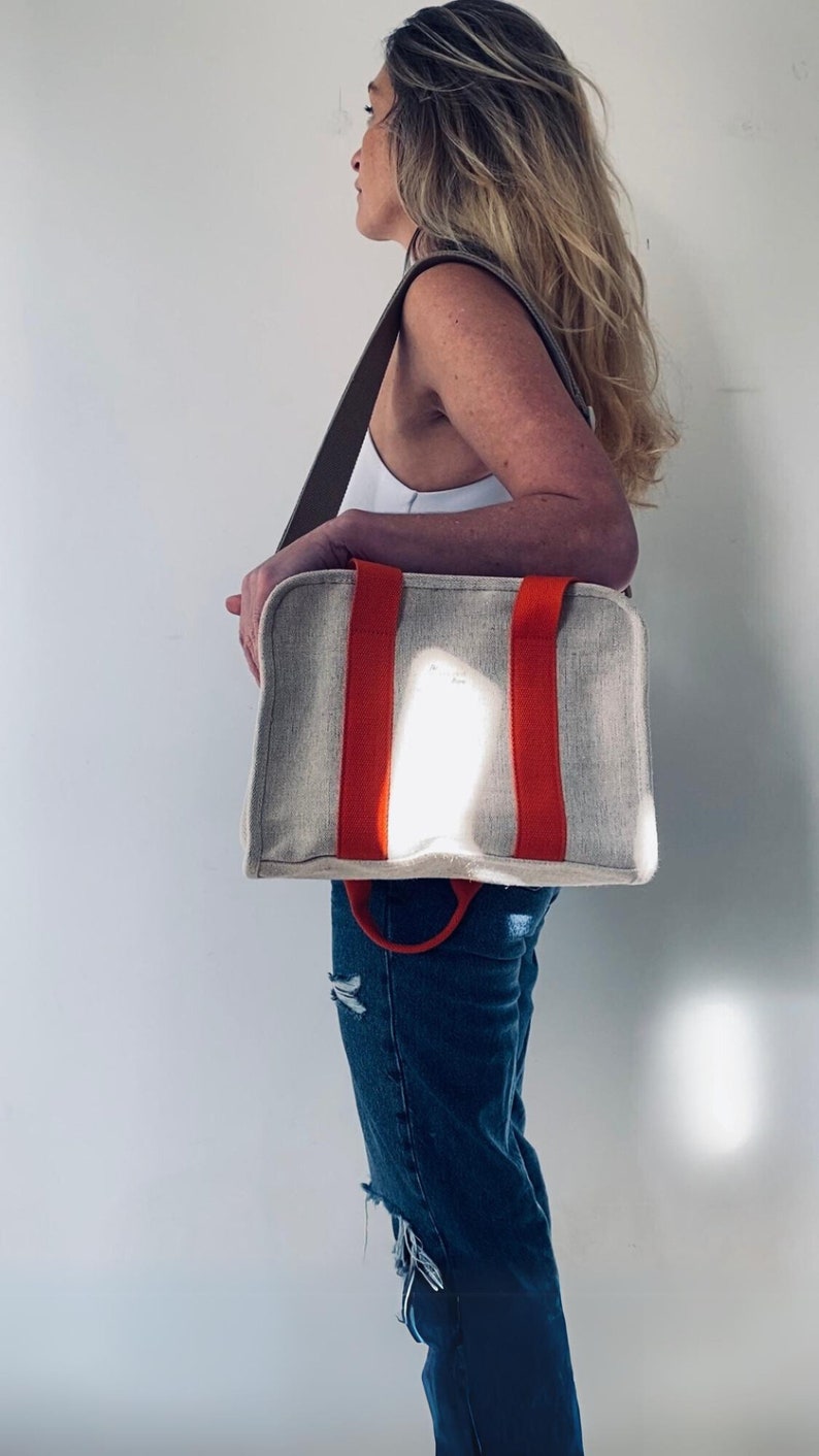 May include: A woman is carrying a beige canvas tote bag with orange straps. The bag has a top handle and a shoulder strap.