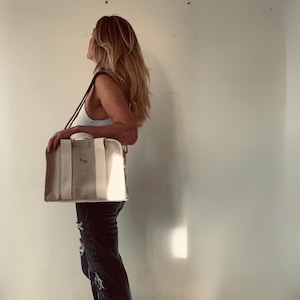 May include: A woman is carrying a beige canvas tote bag with a white leather trim and a brown strap. The bag has a small logo on the front.