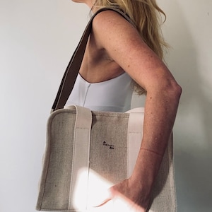 May include: A beige canvas tote bag with a brown leather strap and white handles. The bag has the text "The  Honest  Goods" printed on the front.