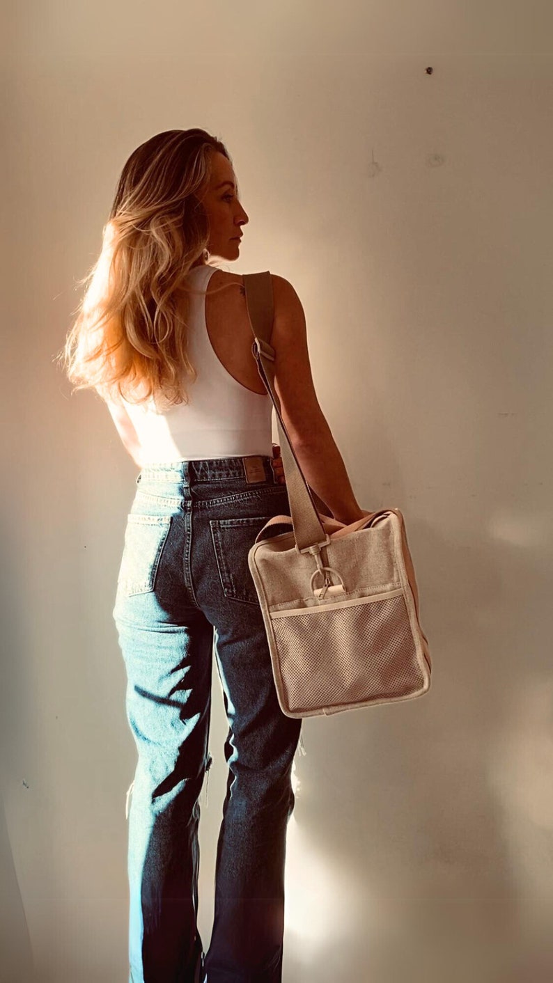 May include: A woman wearing a white tank top and blue jeans is carrying a beige canvas tote bag with a brown strap over her shoulder.