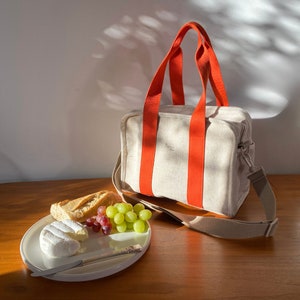 May include: A beige canvas tote bag with orange straps and a brown leather shoulder strap. The bag is sitting on a wooden table next to a plate with a baguette, grapes, and a round cheese.