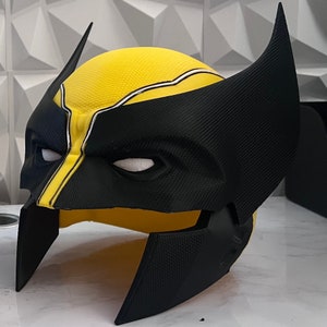 Puede incluir: Una máscara de Wolverine de plástico negro y amarillo con una superficie texturizada. La máscara tiene una franja blanca en relieve en la frente y una máscara de ojos blanca.