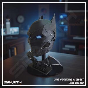 Può includere: Un cappuccio di Batman nero con occhi a LED blu chiaro, esposto su un supporto nero. Il cappuccio ha un aspetto invecchiato e fa parte di un set LED. Il testo "LIGHT WEATHERING W/ LED SET LIGHT BLUE LED" è visibile.