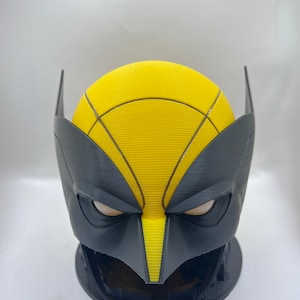 May include: A black and yellow 3D printed mask of a superhero. The mask has a yellow face with black eyes and a black nose. The mask is on a black stand.