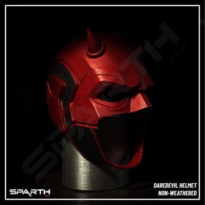 May include: A red Daredevil helmet with a matte finish, featuring two curved horns on top. The helmet has a detailed design with black mesh sections around the eyes and mouth. The text "DAREDEVIL HELMET NON-WEATHERED" is visible.