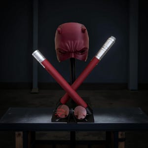 Könnte beinhalten: Eine rote Daredevil-Maske mit Hörnern, zwei rote Schlagstöcke und eine rote Sonnenbrille auf einem schwarzen Ständer.