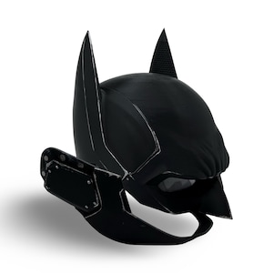 May include: Black Batman mask with pointed ears and a textured surface. The mask features a stylized design with a dark color scheme and a visible eye opening. The mask is designed to be worn and is likely made of a durable material.