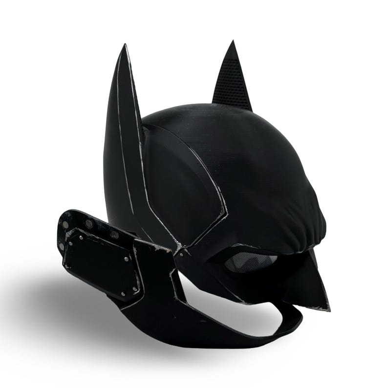 May include: Black Batman mask with pointed ears and a textured surface. The mask features a stylized design with a dark color scheme and a visible eye opening. The mask is designed to be worn and is likely made of a durable material.