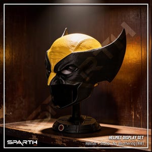 May include: A yellow and black Wolverine helmet replica, displayed on a black stand. The helmet features the character's signature mask design and is part of a helmet display set. The text "HELMET DISPLAY SET" is visible.