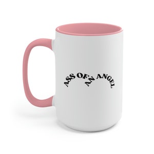 May include: White ceramic mug with a pink rim and handle. The mug has black text that reads "ASS OF AN ANGEL" in a curved arc.