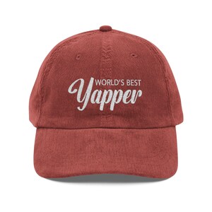 May include: A brick-red corduroy baseball cap with the text "WORLD'S BEST Yapper" embroidered in white script. The cap has a curved peak and an adjustable strap.