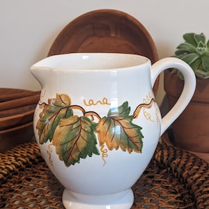 May include: White ceramic pitcher with a vine and leaf design in shades of green and yellow. The pitcher has a rounded body and a handle.