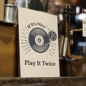 May include: A white sign with black text and a graphic of a record on a turntable. The text reads "If It's Nice! Play It Twice."