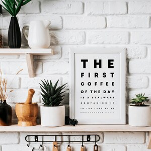 May include: White framed print with black text that reads "THE FIRST COFFEE OF THE DAY IS A STALWART COMPANION IN THE FACE OF AN UNREASONABLE WORLD". The print is on a shelf with a white pitcher, a potted succulent, a wooden mortar and pestle, and a potted plant.