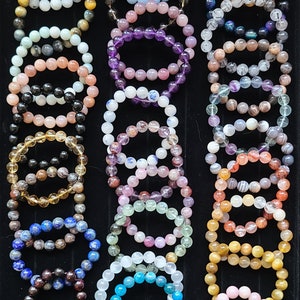 May include: A collection of numerous beaded bracelets arranged in three vertical columns on a black display tray. The bracelets feature various polished gemstones in diverse colors including white, blue, purple, pink, yellow, and brown.