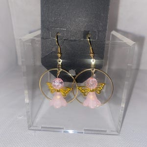 May include: A pair of gold-colored hoop earrings. Each earring features a pink angel charm with gold-colored wings and a pink bead. The earrings are displayed in a clear plastic box against a black background.