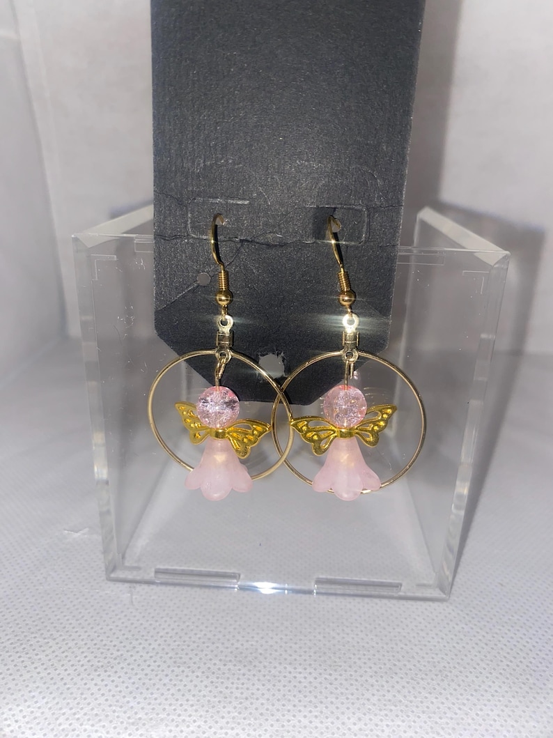 May include: A pair of gold-colored hoop earrings. Each earring features a pink angel charm with gold-colored wings and a pink bead. The earrings are displayed in a clear plastic box against a black background.