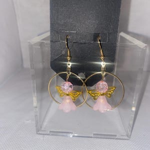 May include: A pair of gold-colored hoop earrings featuring pink angel charms. Each charm has a pink bead head, gold wings, and a pink bell-shaped body. The earrings are displayed on a black card.