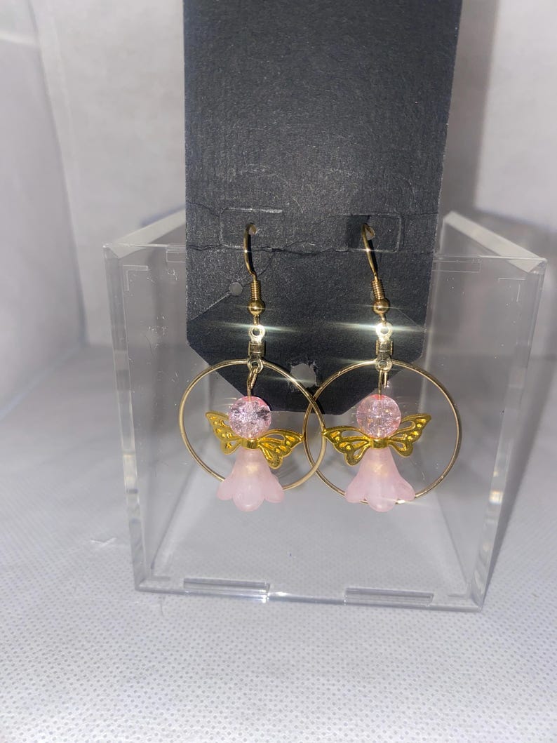 May include: A pair of gold-colored hoop earrings featuring pink angel charms. Each charm has a pink bead head, gold wings, and a pink bell-shaped body. The earrings are displayed on a black card.