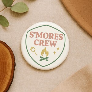 May include: White round button with a green border and the text "S'mores Crew" in pink and yellow. The button also features a small illustration of a marshmallow on a stick, a campfire, and two stars.